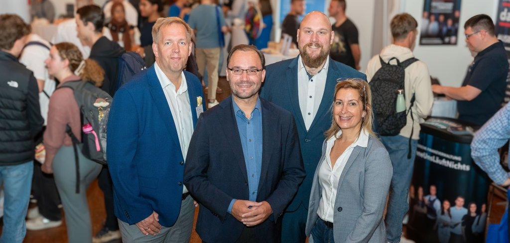 Vier Personen stehen vorne und lächeln in die Kamera. Drei Männer und eine Frau tragen schicke Kleidung. Hinter ihnen sind viele junge Menschen mit Rucksäcken. Sie laufen herum, schauen sich Stände an und sprechen mit anderen. Auf den Ständen gibt es Plakate und Broschüren. Die Veransaltung ist eine Ausbildungsmesse. Hier treffen Firmen und junge Menschen zusammen, die eine Ausbildung suchen.