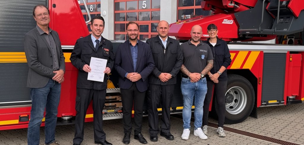 Das Bild zeigt sechs Personen vor einem großen, roten Feuerwehrwagen. Auf dem Fahrzeug steht "Feuerwehr Mühlheim" und die Notrufnummer 112. Ein Mann in schwarzer Feuerwehr-Uniform hält eine Urkunde in der Hand. Er wurde zum stellvertretenden Feuerwehr-Chef ernannt. Neben ihm stehen der Bürgermeister und der Erste Stadtrat von Mühlheim. Alle lächeln und freuen sich über die Ernennung.