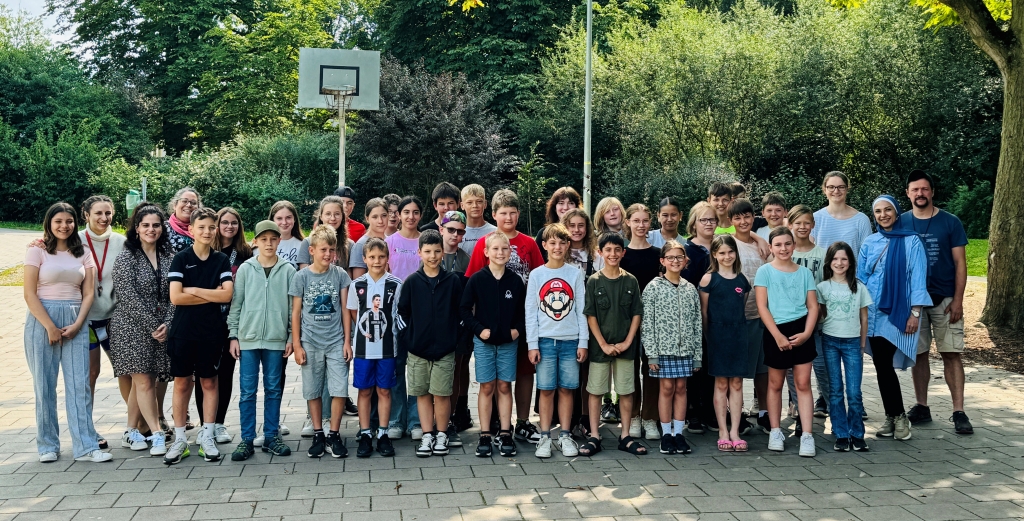 Das Bild zeigt eine Gruppe von Kindern und Betreuern. Sie stehen draußen auf einem Schulhof mit einem Basketballkorb. Alle lächeln und schauen in die Kamera.