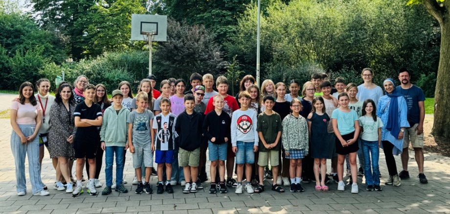 Das Bild zeigt eine Gruppe von Kindern und Betreuern. Sie stehen draußen auf einem Schulhof mit einem Basketballkorb. Alle lächeln und schauen in die Kamera.