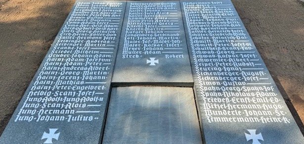 Das Bild zeigt ein Kriegsdenkmal auf einem Friedhof. Das Denkmal besteht aus drei großen, dunklen Steinplatten. Auf den Platten stehen viele Namen in weißer Schrift. Die Stadt Mühlheim hat das Denkmal renoviert. Mitarbeitende vom Friedhof haben dabei geholfen.