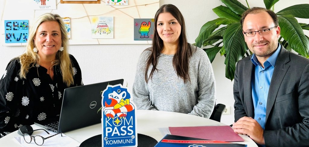 Drei Personen sitzen an einem Tisch. Auf dem Tisch liegt ein Ordner mit einem blauen Logo. Das Logo heißt "Kompass Kommune". Eine Frau übergibt ihre Aufgabe an eine andere Frau. Der Bürgermeister ist auch dabei. Im Hintergrund sind bunte Bilder von Kindern.
