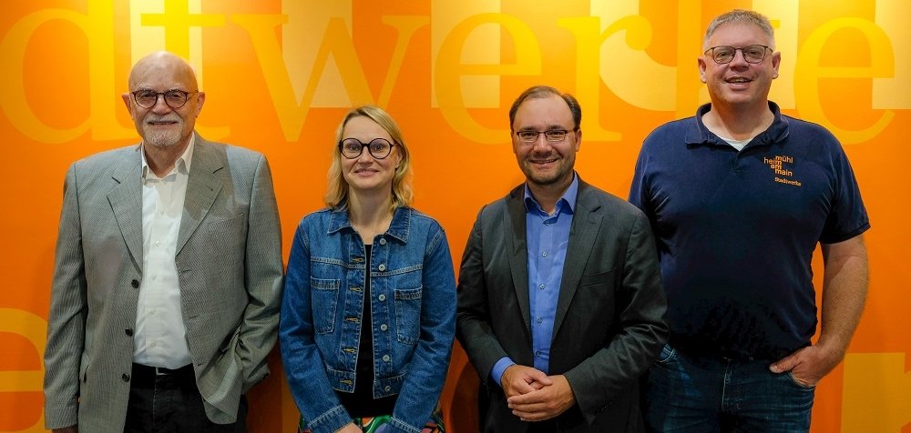 Vier Personen stehen vor einer orangenen Wand. Sie lächeln in die Kamera. Eine Frau ist die neue zusätzliche Chefin der Stadtwerke. Sie fängt am 1. Juli 2024 an. Der Bürgermeister ist auch dabei.