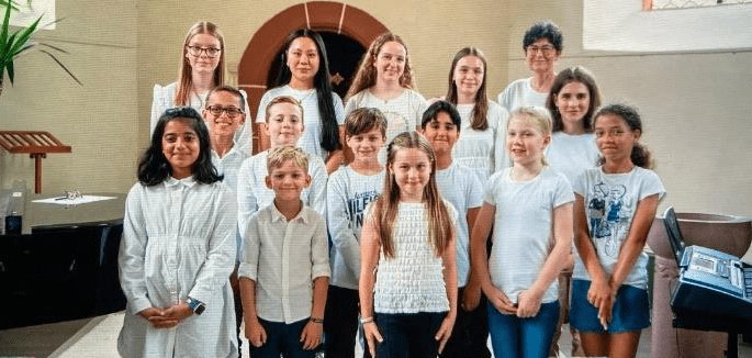 Das Bild zeigt eine Gruppe von Kindern und Jugendlichen in einer Kirche. Alle tragen weiße Kleidung und stehen vor einem Altar mit einem Fenster. Sie haben ein Konzert der Musikschule Mühlheim gegeben. Die Kinder haben Klavier gespielt und gesungen.