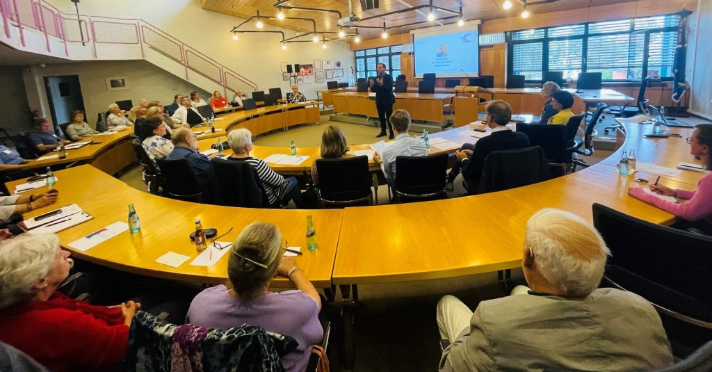 Viele Menschen sitzen an einem großen runden Tisch in einem großen Raum. Vorne steht Bürgermeister Dr. Alexander Krey im Anzug. Er spricht zu den Menschen und hält eine Präsentation. Auf den Tischen liegen Papiere, Stifte und Wasserflaschen. Der Raum hat große Fenster, durch die Licht hereinkommt.