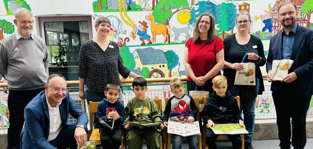 Auf dem Bild sind vier Kinder und sechs Erwachsene in einer Bücherei zu sehen, Die Kinder sitzen auf Stühlen und halten Bücher in den Händen. Sie schauen in die Bücher und lächeln. Die Erwachsenen stehen hinter ihnen und freuen sich. Zwei Männer, der Bürgermeister und der Landrat, halten kleine Stofftaschen mit Büchern. Im Hintergrund ist eine bunte Wand mit Bildern von Märchenfiguren.