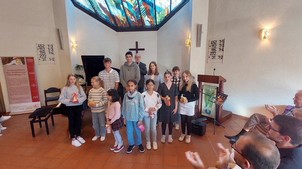 Eine Gruppe von Kindern und Jugendlichen steht in einer Kirche. Sie halten kleine Geschenke in den Händen. Hinter ihnen ist ein buntes Kirchenfenster und ein Kreuz. Vorne klatschen einige Erwachsene.