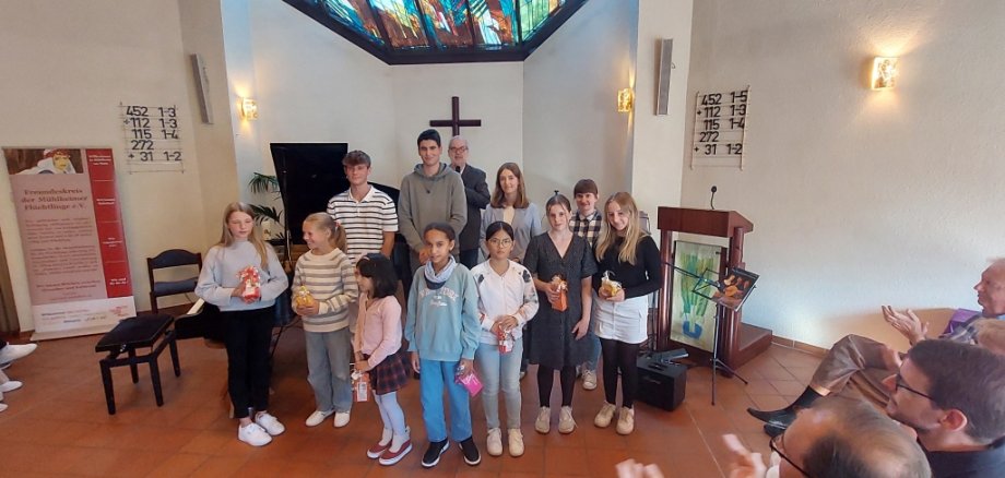 Eine Gruppe von Kindern und Jugendlichen steht in einer Kirche. Sie halten kleine Geschenke in den Händen. Hinter ihnen ist ein buntes Kirchenfenster und ein Kreuz. Vorne klatschen einige Erwachsene.