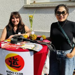 Zwei Frauen stehen an einem Infostand des Budo Clubs Mühlheim. Auf dem Tisch liegt ein schwarzer Judo-Gürtel, Flyer und kleine Kürbisse als Dekoration. Ein großes Plakat mit dem Logo des Vereins hängt am Tisch. Im Hintergrund sieht man ein Gebäude mit einem Fenster. Die Veranstaltung findet draußen statt.