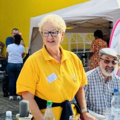 Eine Frau in einem gelben T-Shirt steht an der langen Tafel. Sie heißt Erika Sickenberger und arbeitet für die Stadt Mühlheim. Sie kümmert sich um ältere Menschen. Sie ist heute hier, weil der Bürgermeister nicht kommen konnte. Sie vertritt ihn bei dieser Veranstaltung. Hinter ihr ist ein Zelt mit einem Banner. Sie spricht mit den anderen Menschen an der Tafel.