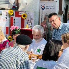Mehrere Menschen sitzen draußen an einem langen Tisch. Auf dem Tisch stehen Teller, Gläser und Wasserflaschen. Im Hintergrund gibt es rote und weiße Luftballons. Auch Sonnenblumen und Plakate sind zu sehen. Ein Mann mit Brille und kariertem Hemd steht und spricht mit den Leuten am Tisch. Hinter ihm hängt ein großes Plakat mit dem Logo der "Clown Doktoren".