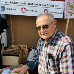 Ein Mann mit Sonnenbrille und kariertem Hemd sitzt an einem Stand des Geschichtsvereins. Er hat Infomaterial in der Hand. Hinter ihm hängt ein großes Schild mit den Wappen der Stadtteile von Mühlheim. Neben ihm stehen Taschen und Flyer.