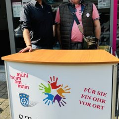 Zwei Männer stehen hinter einem Infostand. Auf dem Stand steht "Stadtteil-Kümmerer". Sie helfen Menschen im Stadtteil. Sie hören zu und kümmern sich um Probleme.