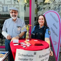 Ein älterer Mann mit Mütze und eine Polizistin stehen an einem Tisch. Der Mann ist Sicherheitsberater für Senioren. Die Polizistin ist eine Schutzfrau vor Ort. Sie geben Tipps, wie man sicher leben kann.
