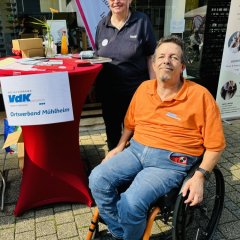 Eine Frau und ein Mann im Rollstuhl stehen an einem Tisch. Der Tisch gehört zum VdK. Der VdK hilft Menschen mit Behinderungen. Es gibt Infomaterialien und Getränke.