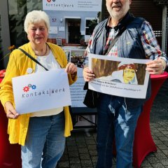 Eine Frau und ein Mann stehen vor einem Gebäude mit großen Fenstern. Sie halten Schilder mit der Aufschrift "Kontakt-Werk" und "Wir bauen Brücken". Hinter ihnen ist ein Infostand mit weiteren Informationen. Der Mann trägt eine Weste und eine Tasche über der Schulter. Die Frau trägt eine gelbe Jacke und lächelt in die Kamera.