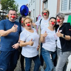 Eine Gruppe von Menschen trägt weiße T-Shirts und rote Clownsnasen. Sie lachen und zeigen mit den Daumen nach oben. Hinter ihnen sieht man Infostände und Besucher. Die Gruppe gehört zum Roten Kreuz und möchte mit Humor helfen.