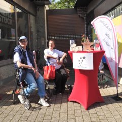 Zwei Frauen sitzen an einem Infostand auf der Straße. Beide nutzen Rollatoren als Sitzmöglichkeit. Sie gehören zu einem Verein, der sich für Menschen mit Multipler Sklerose einsetzt. Vor ihnen steht ein roter Stehtisch mit Flyern und Informationen über ihre Arbeit. Hinter ihnen ist ein Banner mit dem Namen "MainSterne" zu sehen.