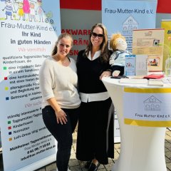 Zwei Frauen stehen vor einem weißen Stehtisch. Sie lächeln in die Kamera. Hinter ihnen sind große Plakate mit bunten Zeichnungen von Kindern. Auf dem Tisch liegen Flyer, Broschüren und ein kleines Kuscheltier. Die Frauen stellen Informationen für Eltern und Kinder bereit.