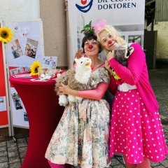 Zwei Frauen sind als Clowns verkleidet. Sie haben bunte Kleidung und rote Nasen. Sie stehen vor einem Stand mit einem Plakat. Sie wollen kranken Menschen Freude bringen.