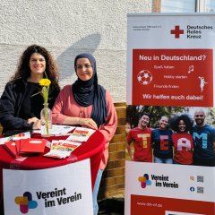 Zwei Frauen stehen an einem Infostand mit Flyern und Informationen des Deutschen Roten Kreuzes. Ein großes Plakat mit den Worten "Neu in Deutschland?" informiert über Angebote für Zugewanderte. Im Hintergrund ist eine weiße Wand mit Fenstern zu sehen.