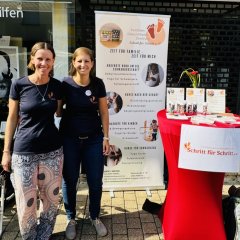 Zwei Frauen stehen vor einem Stand mit einem roten Tisch. Sie lächeln in die Kamera. Hinter ihnen ist ein Plakat mit Informationen über Kurse für Familien. Neben dem Tisch liegen Flyer und Broschüren. Es ist sonnig und sie stehen vor einem Geschäft.