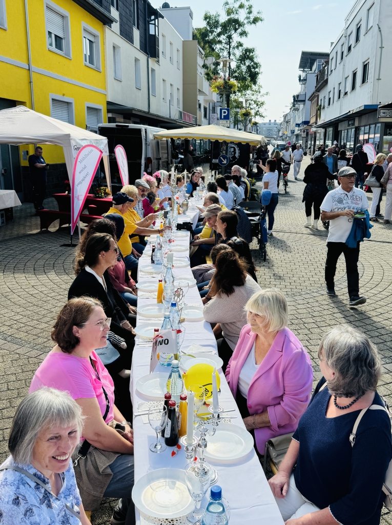 Viele Menschen sitzen an einer langen Tafel auf der Straße. Sie essen und trinken gemeinsam. Die Straße ist festlich geschmückt. Es gibt auch Stände mit Informationen über das Ehrenamt. Es ist ein schöner Tag mit Sonne.