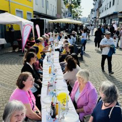 Viele Menschen sitzen an einer langen Tafel auf der Straße. Sie essen und trinken gemeinsam. Die Straße ist festlich geschmückt. Es gibt auch Stände mit Informationen über das Ehrenamt. Es ist ein schöner Tag mit Sonne.