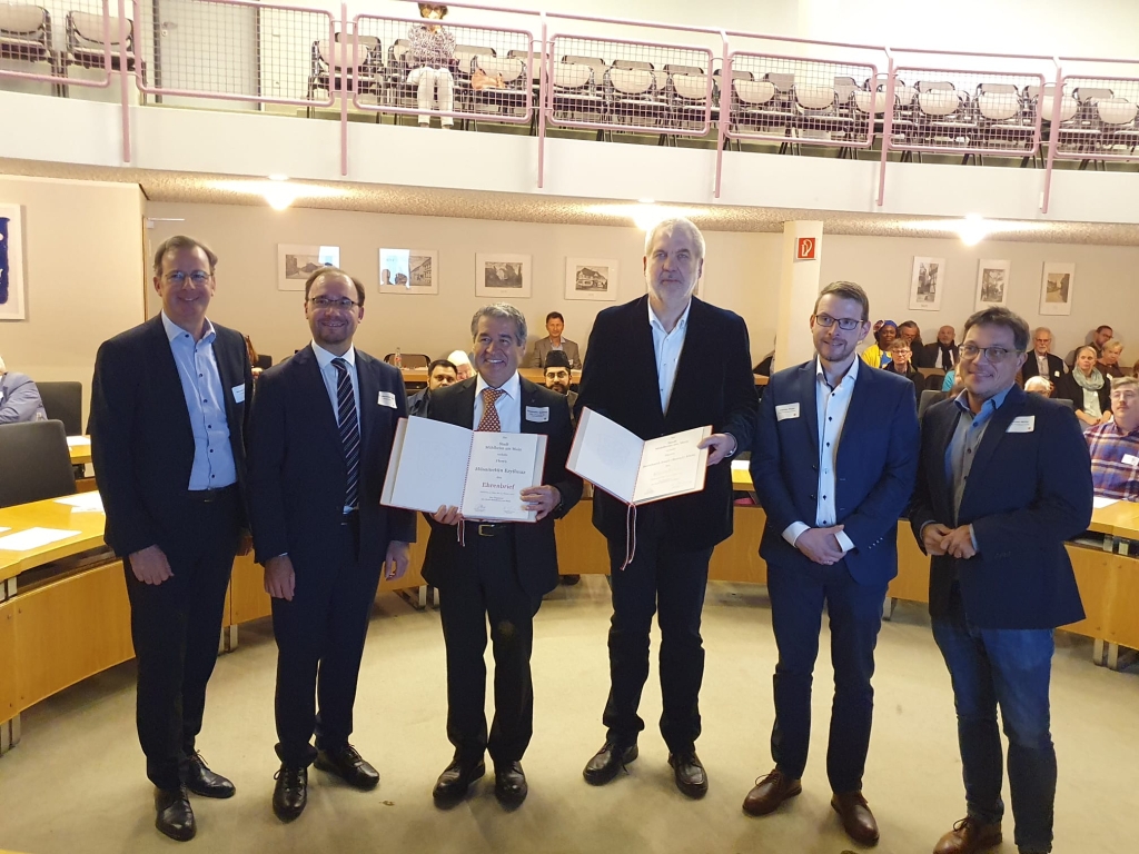 Sechs Männer stehen in einem großen Saal. Zwei Männer in der Mitte halten große Urkunden in den Händen. Sie lächeln und schauen in die Kamera. Im Hintergrund sitzen viele Menschen und schauen zu. Einige sitzen auf einer Empore oben.