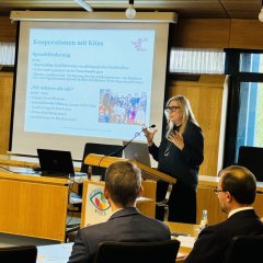 Die Leiterin der Abteilung Jugend und Soziales steht vorne im Raum und hält einen Vortrag. Sie trägt eine Brille und gestikuliert mit den Händen. Hinter ihr sieht man eine große Leinwand mit einer Präsentation. Auf der Leinwand steht "Kooperationen mit Kitas".