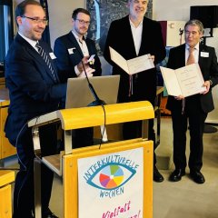 Bürgermeister Dr. Alexander Krey spricht am Rednerpult. Er hält eine kleine Medaille in der Hand und lächelt. Drei Männer stehen neben ihm und halten große Urkunden. Alle tragen einen Anzug. Hinter ihnen hängt eine große Karte von Mühlheim. Auf dem Rednerpult steht ein Schild mit der Aufschrift "Interkulturelle Wochen".