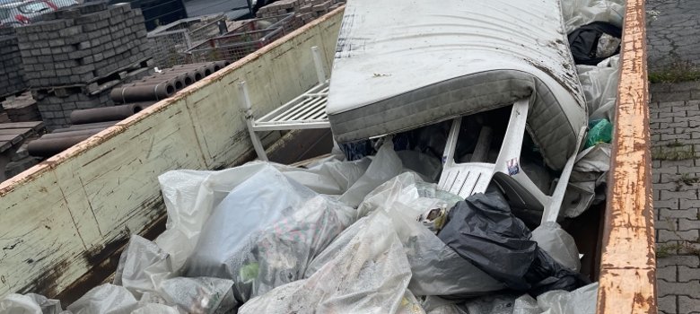 Ein großer Container ist voller Müll. Darin sind Plastiktüten, eine alte Matratze und Metallteile. Im Hintergrund sind Gebäude und Autos zu sehen.