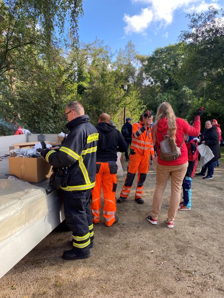 Mehrere Menschen stehen auf einem Platz im Grünen. Einige tragen Feuerwehr- oder Arbeitskleidung. Ein Tisch mit Kartons steht bereit. Eine Frau in roter Jacke spricht mit den Helfern.
