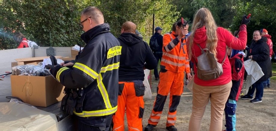 Mehrere Menschen stehen auf einem Platz im Grünen. Einige tragen Feuerwehr- oder Arbeitskleidung. Ein Tisch mit Kartons steht bereit. Eine Frau in roter Jacke spricht mit den Helfern.