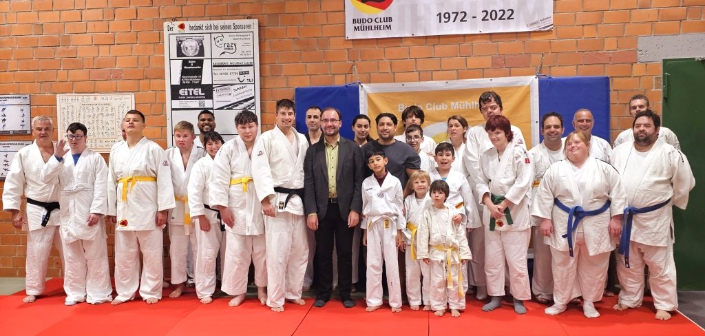 Eine große Gruppe steht auf einer roten Matte. Alle tragen weiße Judo-Anzüge mit farbigen Gürteln. In der Mitte steht der Bürgermeister in einem Anzug. Hinter ihnen hängt ein großes Banner mit der Aufschrift: "50 Jahre Budo Sport in Mühlheim". Alle schauen in die Kamera und lächeln.