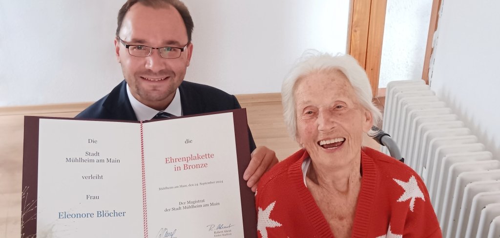 Bürgermeister Dr. Alexander Krey sitzt neben einer älteren Frau. Er hält eine große Urkunde mit ihrem Namen. Die Frau trägt einen roten Pullover mit weißen Sternen. Sie lacht. In ihren Händen hält sie eine Ehrenplakette und eine kleine Medaille.