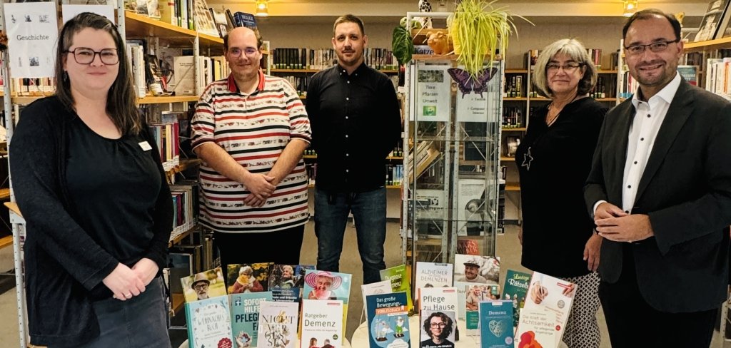 Fünf Menschen stehen in einer Bibliothek. Vor ihnen ist ein Tisch mit vielen Büchern und Spielen zum Thema Demenz. Rechts steht Bürgermeister Dr. Alexander Krey. Alle lächeln in die Kamera.