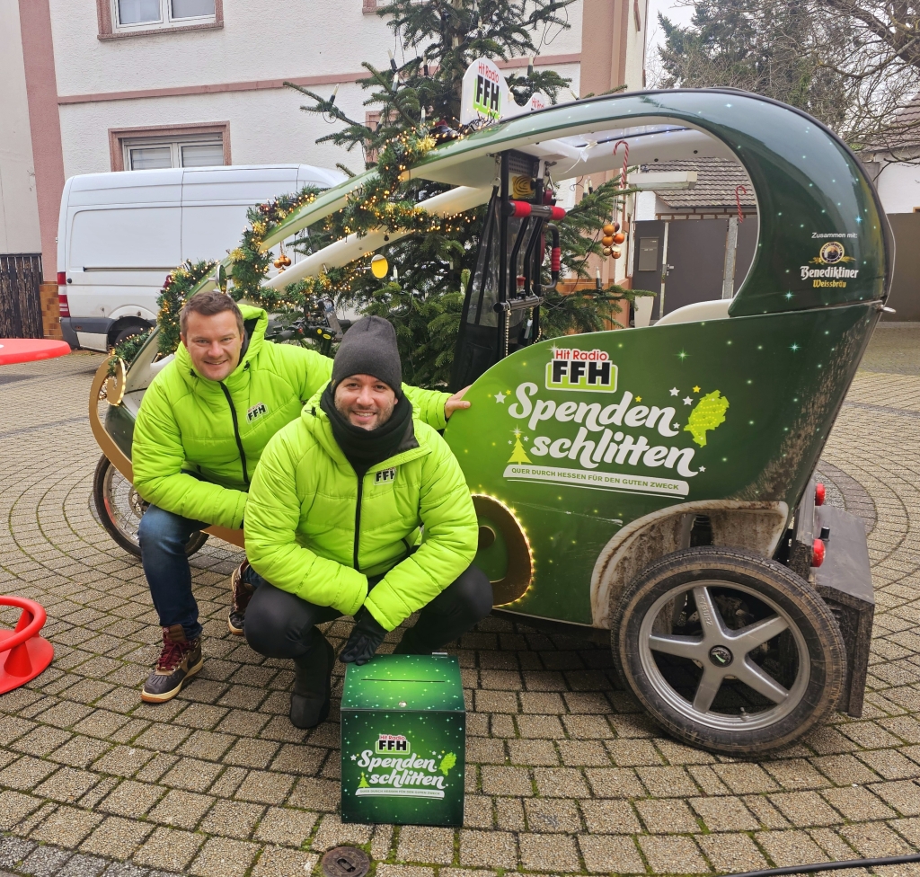 Zwei Männer sitzen vor einem kleinen, grünen Auto. Auf dem Auto steht "FFH Spendenschlitten". Die Männer tragen grüne Jacken. Sie lachen in die Kamera. Einer hält eine grüne Spenden-Box. Hinter ihnen steht ein geschmückter Weihnachtsbaum.