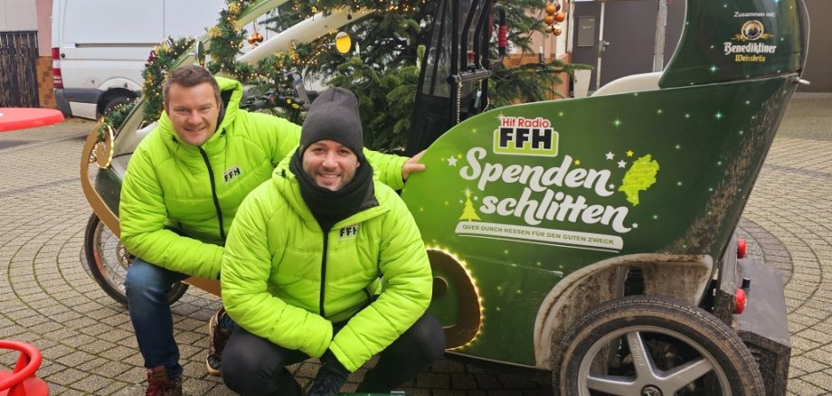 Zwei Männer sitzen vor einem kleinen, grünen Auto. Auf dem Auto steht "FFH Spendenschlitten". Die Männer tragen grüne Jacken. Sie lachen in die Kamera. Einer hält eine grüne Spenden-Box. Hinter ihnen steht ein geschmückter Weihnachtsbaum.
