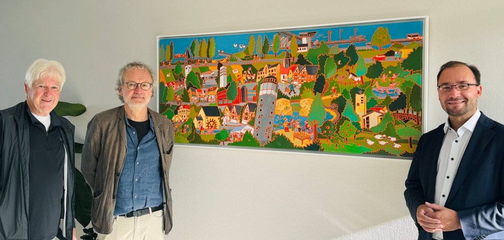 Drei Männer stehen vor einem bunten Bild an einer weißen Wand. Das Bild zeigt viele Häuser, Bäume und bekannte Orte aus Mühlheim. Rechts steht Bürgermeister Dr. Alexander Krey. In der Mitte steht Künstler Klaus Puth, der das Bild gemalt hat. Alle lächeln in die Kamera.