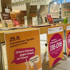Ein Tisch mit vielen Plakaten und Flyern zum Thema "Gewalt gegen Frauen". Auf den Plakaten steht eine Telefonnummer für Hilfe: 116 016. Im Hintergrund hängen Schilder mit Botschaften für Frauenrechte.