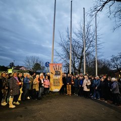 Viele Menschen stehen draußen vor einer Fahne mit der Aufschrift "Stopp Gewalt gegen Frauen". Die Gruppe hält Kerzen und zeigt Unterstützung gegen Gewalt.