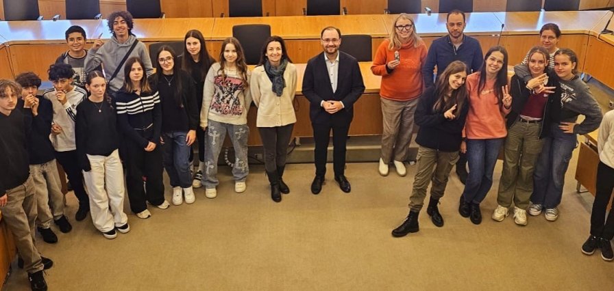 Eine Gruppe von Jugendlichen und Erwachsenen steht in einem großen Saal mit vielen Tischen und Stühlen. In der Mitte steht Bürgermeister Dr. Alexander Krey. Die Jugendlichen lächeln. Sie tragen lockere Kleidung und machen fröhliche Gesten.