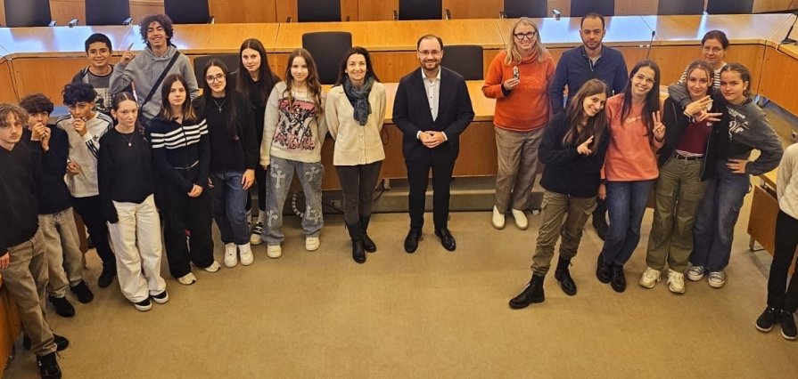 Eine Gruppe von Jugendlichen und Erwachsenen steht in einem großen Saal mit vielen Tischen und Stühlen. In der Mitte steht Bürgermeister Dr. Alexander Krey. Die Jugendlichen lächeln. Sie tragen lockere Kleidung und machen fröhliche Gesten.