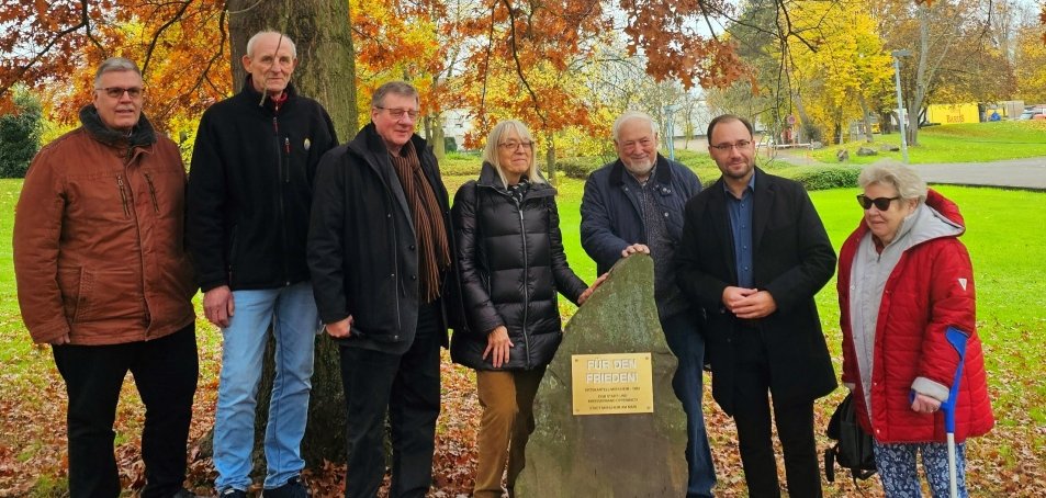 Sieben Menschen stehen vor einem großen Baum mit braunen Blättern. In der Mitte ist ein Gedenkstein mit einem goldenen Schild. Rechts steht Bürgermeister Dr. Alexander Krey. Die Gruppe trägt warme Kleidung und schaut in die Kamera.