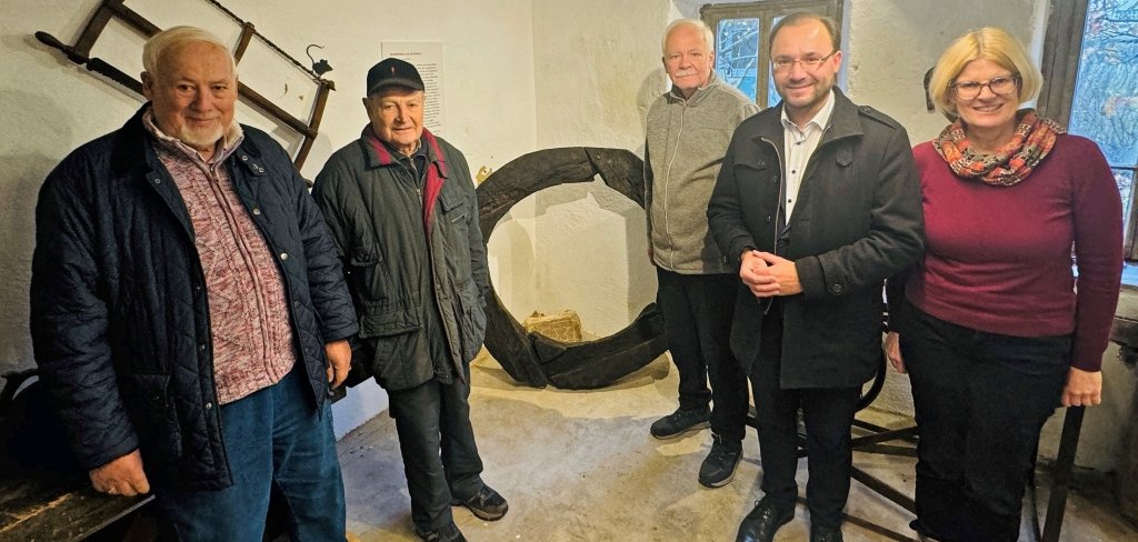 Fünf Personen stehen in einem kleinen Raum. Sie schauen in die Kamera. In der Mitte ist ein großes, schwarzes Holzstück. Das Holzstück ist alt. Es gehört zu einem Brunnen,