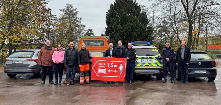 Eine Gruppe von Menschen steht vor mehreren Autos. Sie halten ein rotes Schild mit der Aufschrift "mindestens 1,5 Meter". Die Menschen tragen warme Kleidung. Einige haben Arbeitskleidung an. Im Hintergrund sind Bäume und ein grauer Himmel.