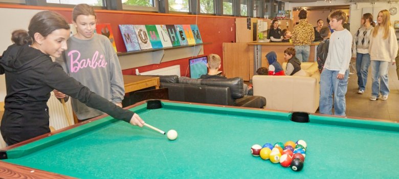 Zwei Mädchen spielen Billard. Im Hintergrund sind Sofas und bunte Bilder an der Wand.