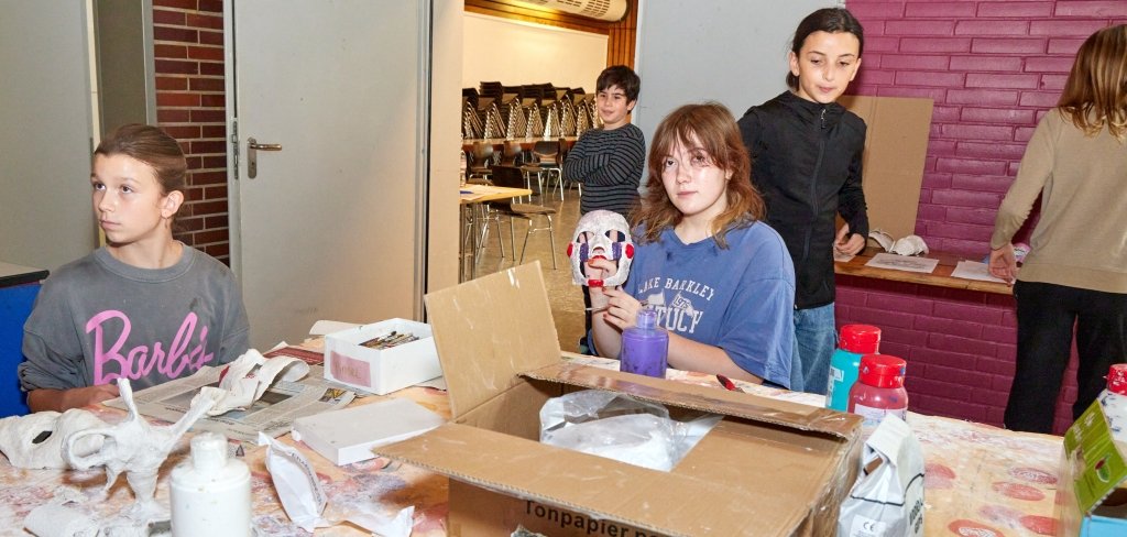 Kinder sitzen an einem Tisch und basteln. Ein Mädchen hält eine bunte Maske hoch. Auf dem Tisch liegen Scheren, Farben und Papier.