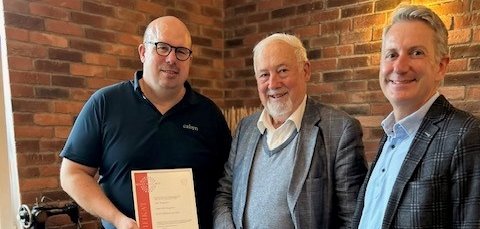 Drei Männer stehen in einem Raum mit Backsteinwand. Einer hält eine Urkunde in der Hand. Alle lächeln und schauen in die Kamera. Im Hintergrund steht eine alte Nähmaschine.